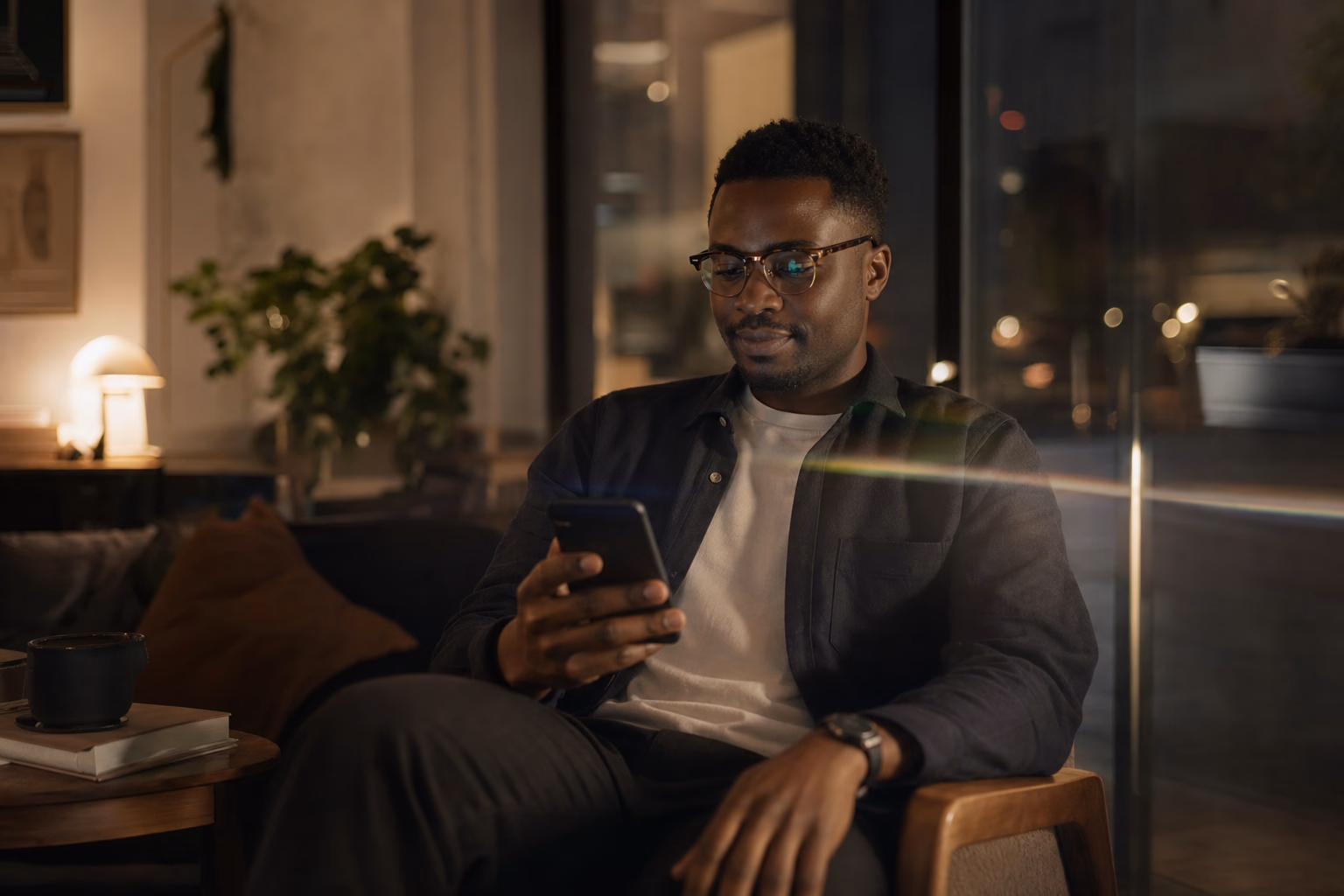 A man sits in a calm, warmly lit room at night checking his phone, relaxed and well-connected